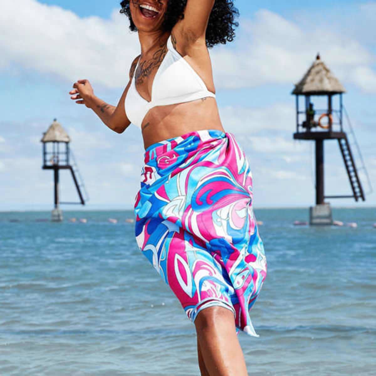 A vibrant blue and pink beach towel features a swirling pattern, held by a woman in a bikini by the water.