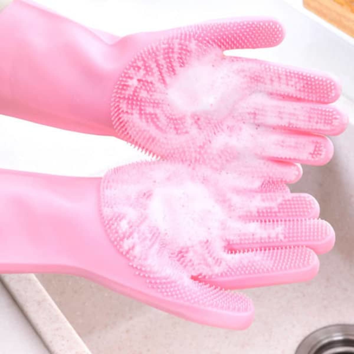 Pink silicone dishwashing gloves with textured palms, covered in soapy suds, held over a sink.