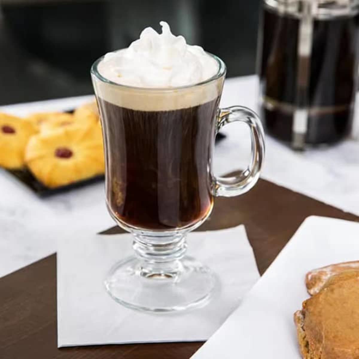 A glass Irish coffee mug filled with dark coffee topped with whipped cream, accompanied by pastries on a table setting.