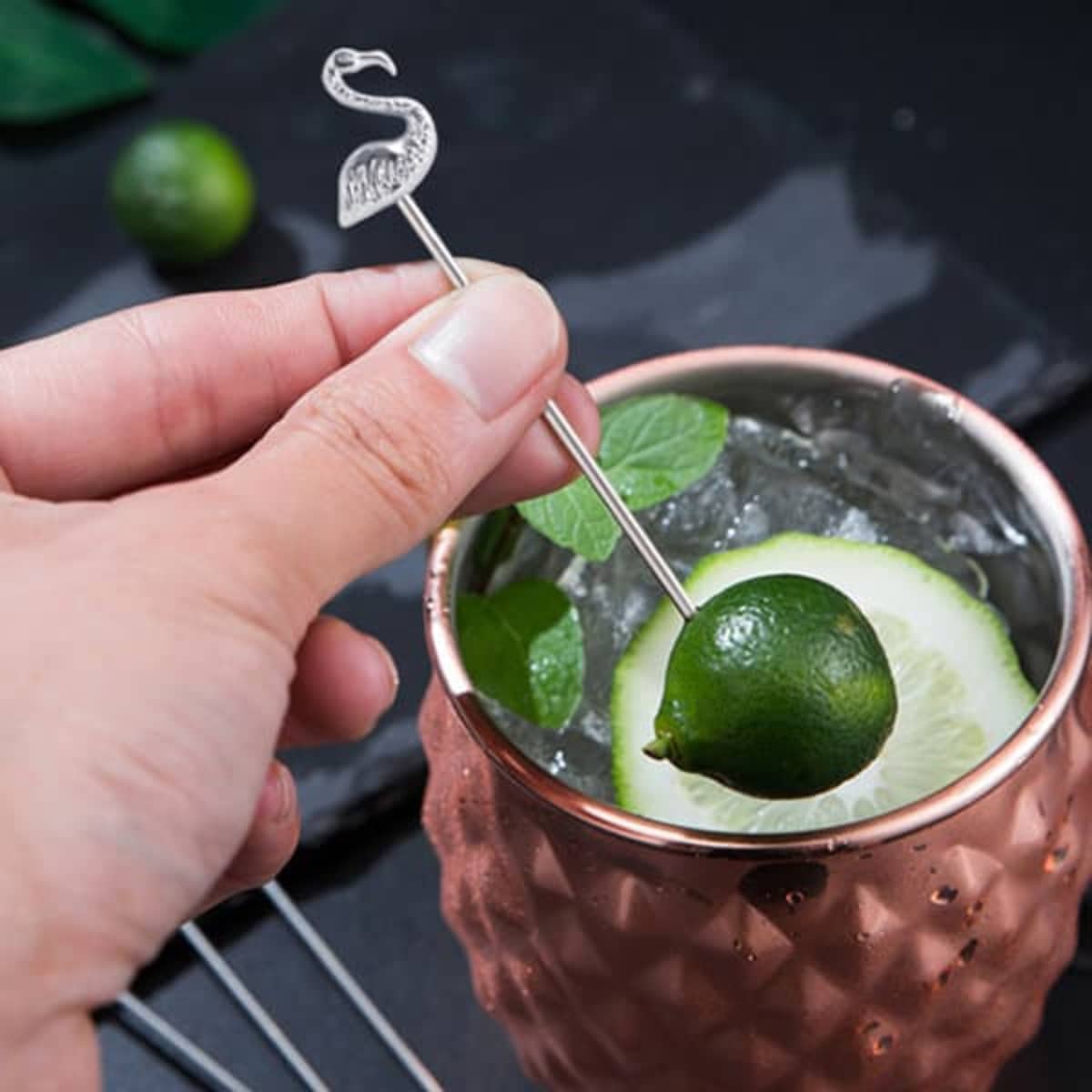 A stainless steel cocktail pick with a flamingo design is poised above a cocktail garnished with lime and cucumber.