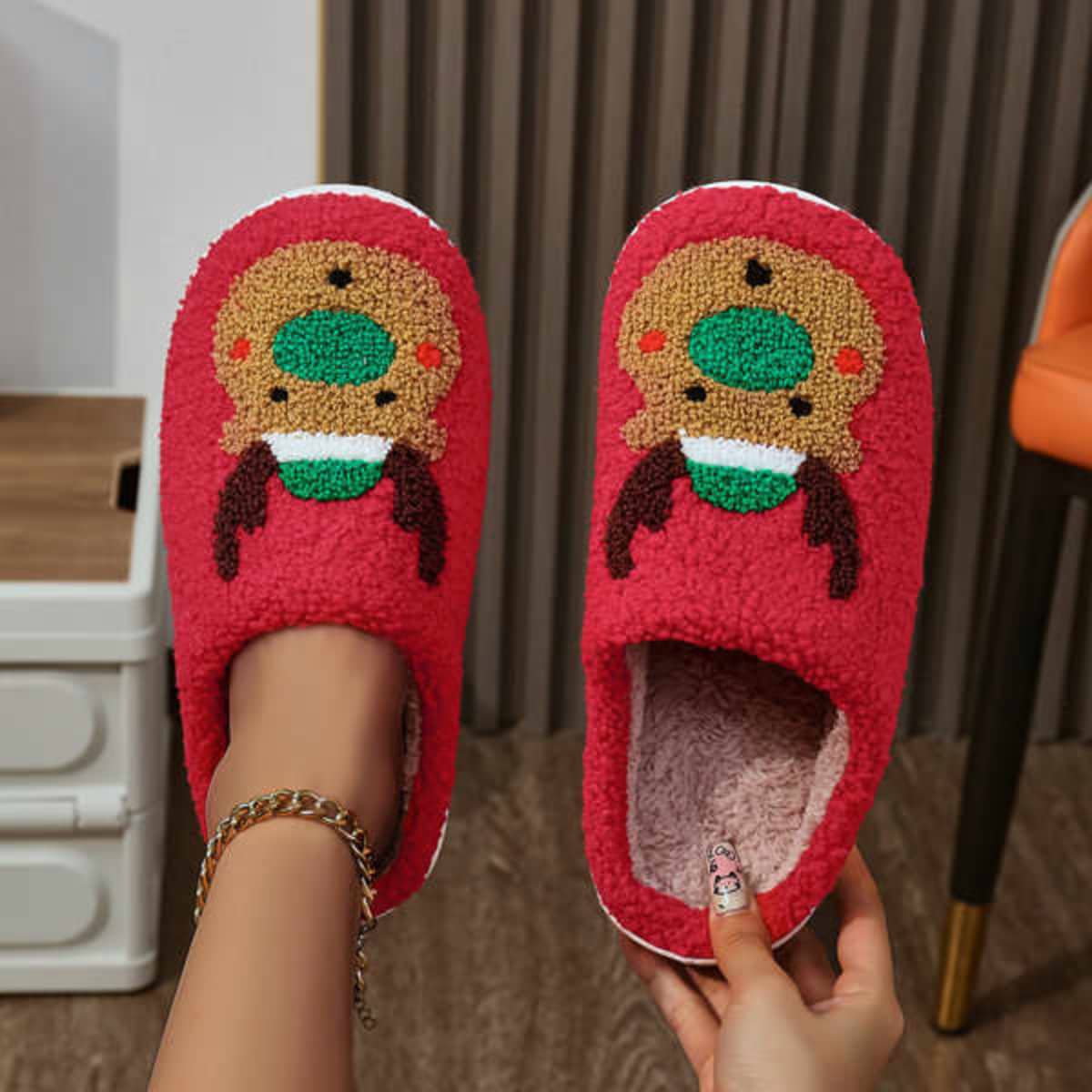 Red fuzzy slippers featuring a reindeer design with green and brown accents, displayed with a person's hands.