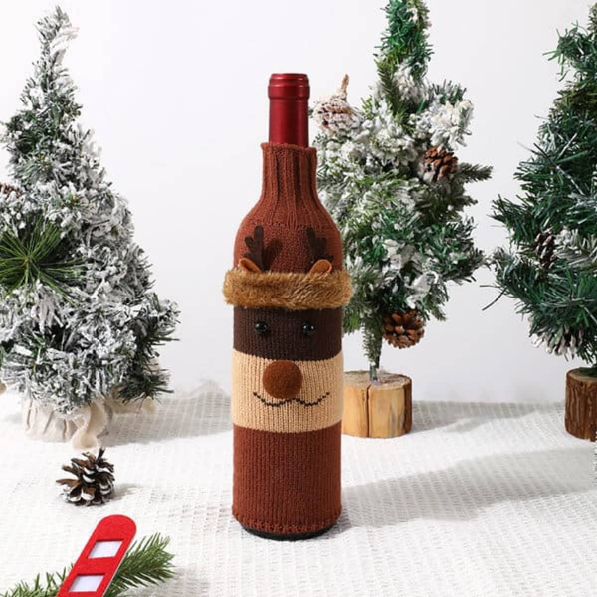 A drink bottle covered in a brown knitted sleeve with a reindeer design, set among miniature Christmas trees.