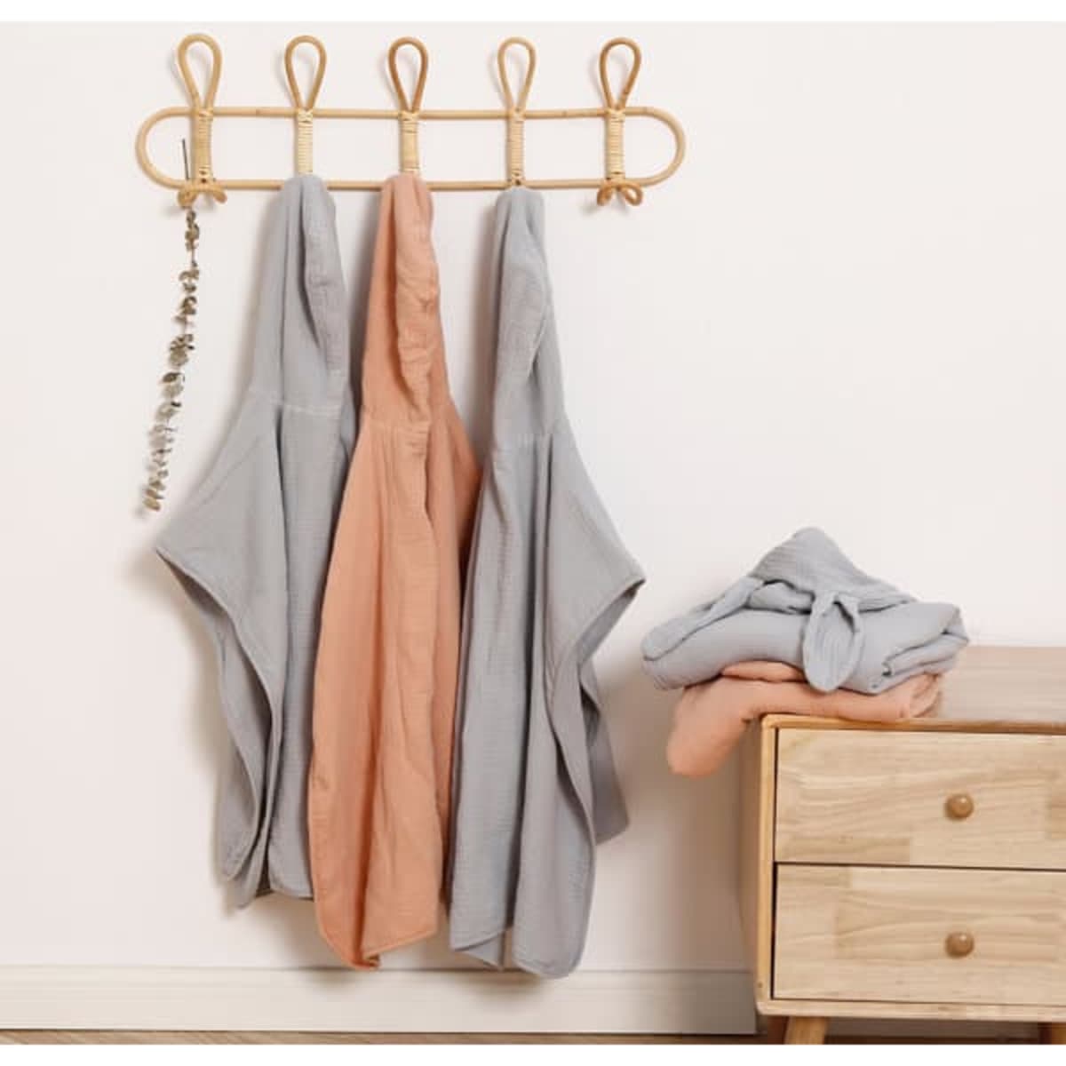 A set of baby hooded poncho towels in grey and peach hanging on a wall, with a folded towel beside a wooden dresser.
