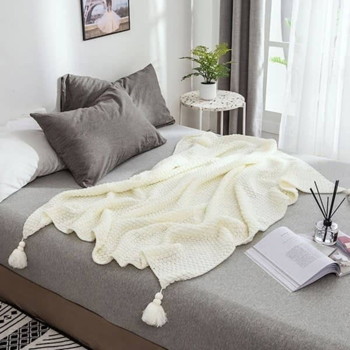 A soft cream Knit Tassel Throw Blanket drapes over a bed with grey pillows, set in a bright, airy room.