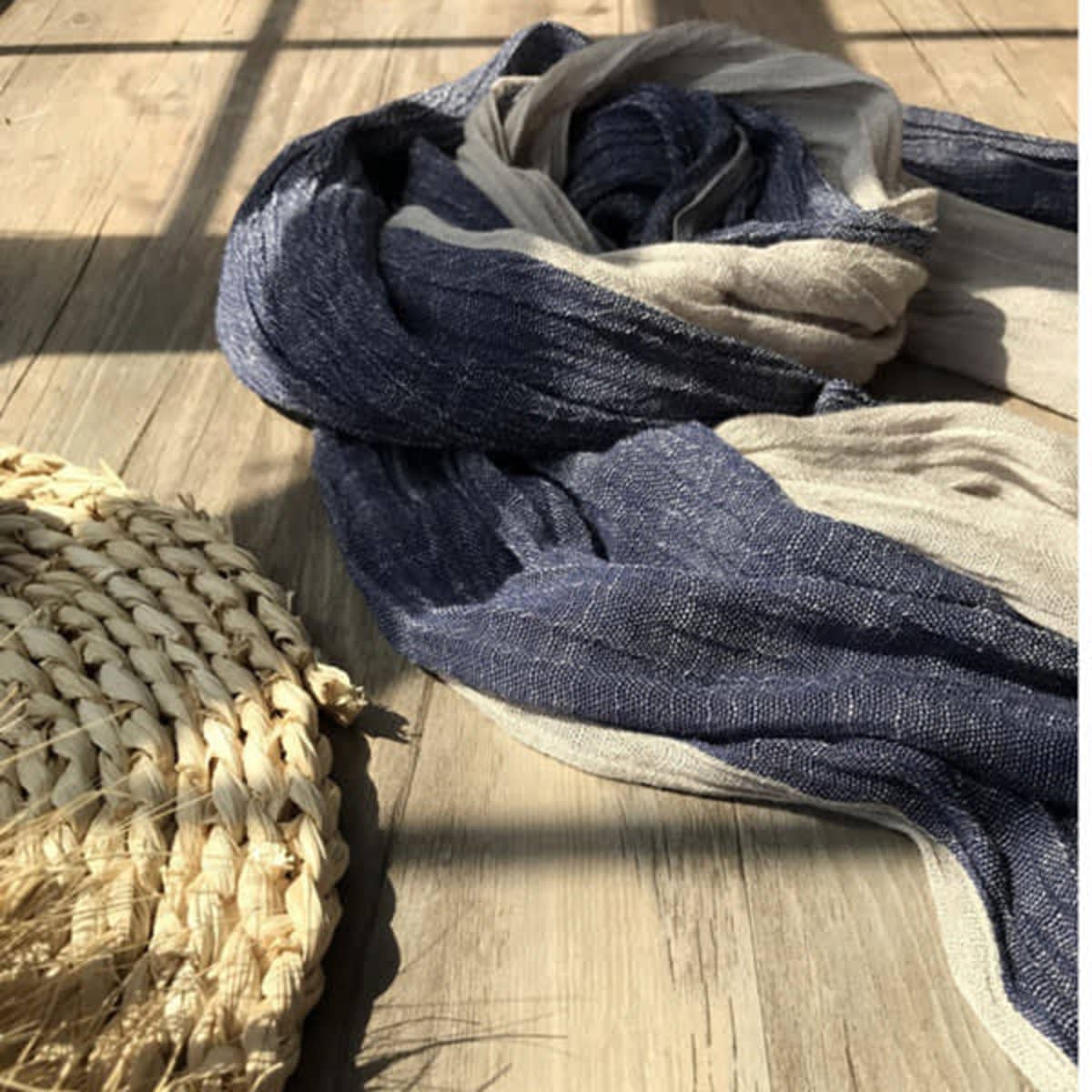 A navy and beige soft shawl wrap scarf lies on a wooden surface next to a woven basket. The scarf has tassels.