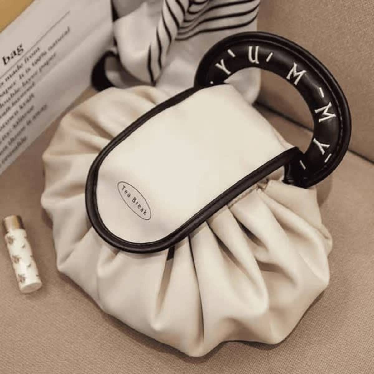 A drawstring makeup bag in cream with a black handle featuring lettering and a rounded design.