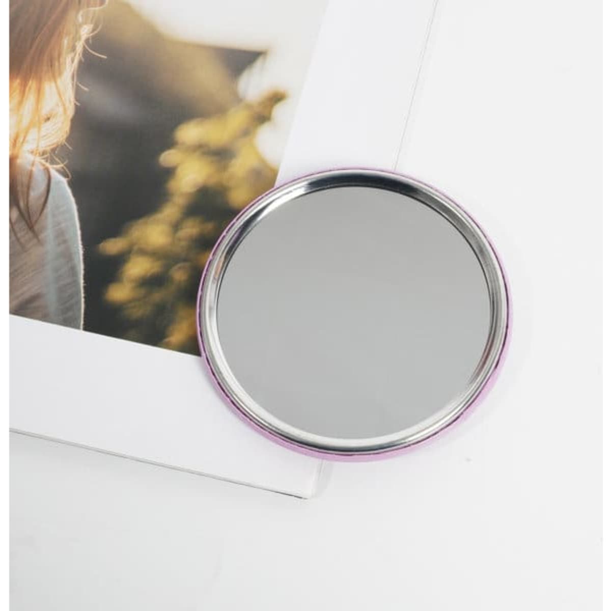 A circular tinplate pocket mirror with a silver reflective surface and a pink border, placed next to a photograph.