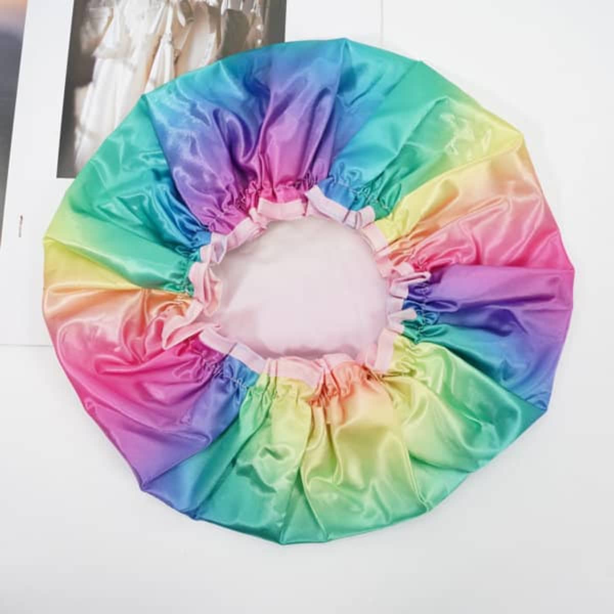 A custom shower cap featuring a vibrant rainbow gradient of pink, purple, and green shades, with an elasticated edge and a logo.