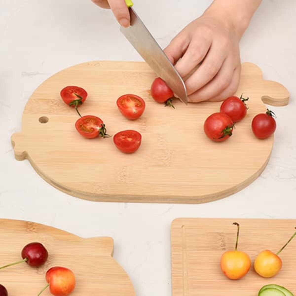 A bamboo cutting board in a playful shape, with cherry tomatoes being chopped and other fruits beside it.