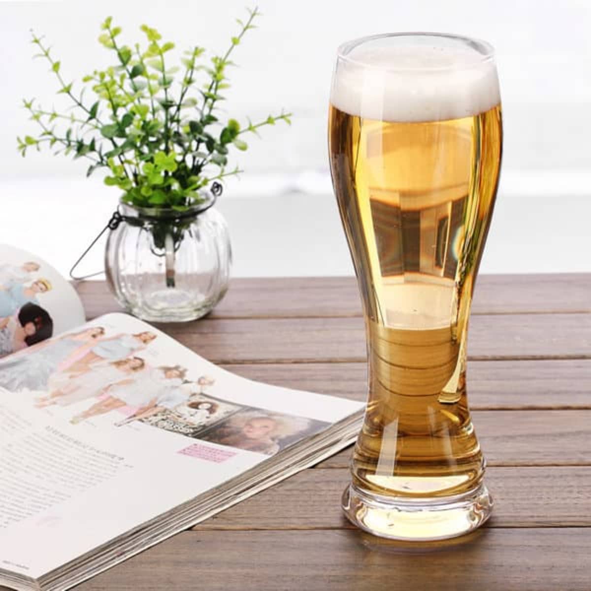 A 22oz. wheat beer glass filled with golden beer and topped with foam, placed on a wooden table beside a magazine and a small plant.