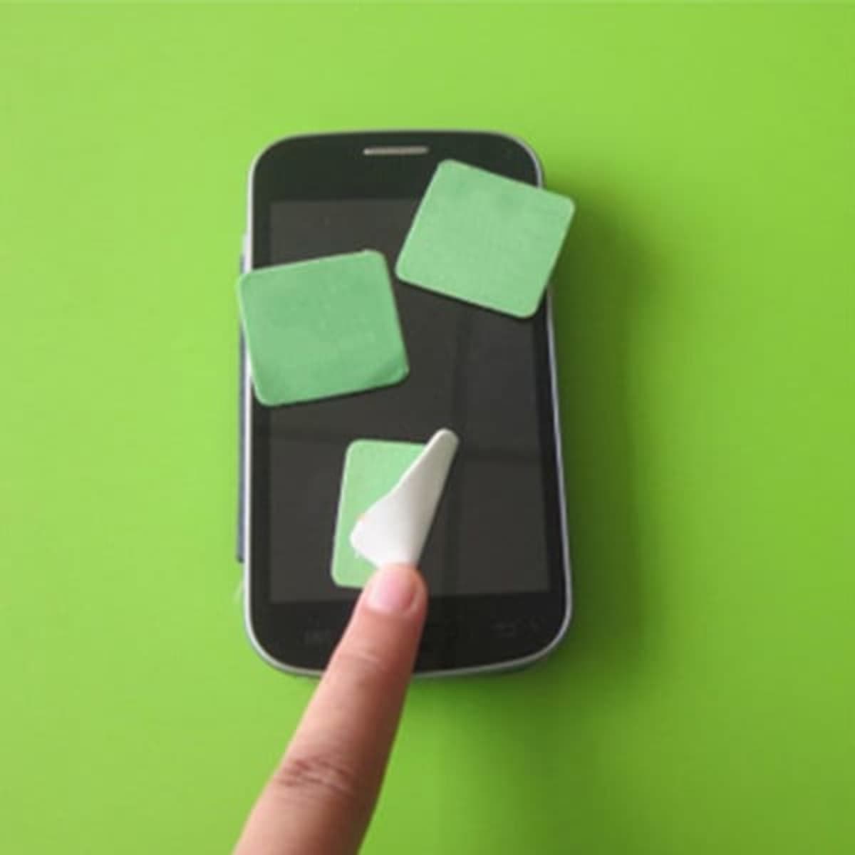 A smartphone on a green background with three green sticky screen cleaners and a finger applying one.