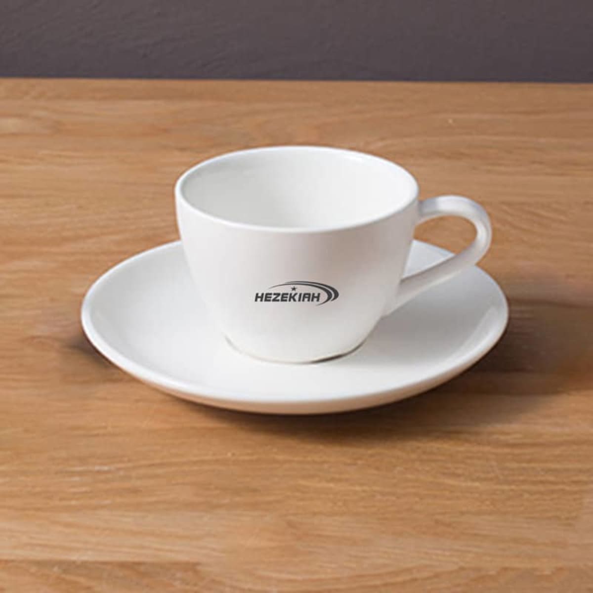 A white porcelain coffee cup with a matching saucer, featuring a simple logo, resting on a wooden surface.