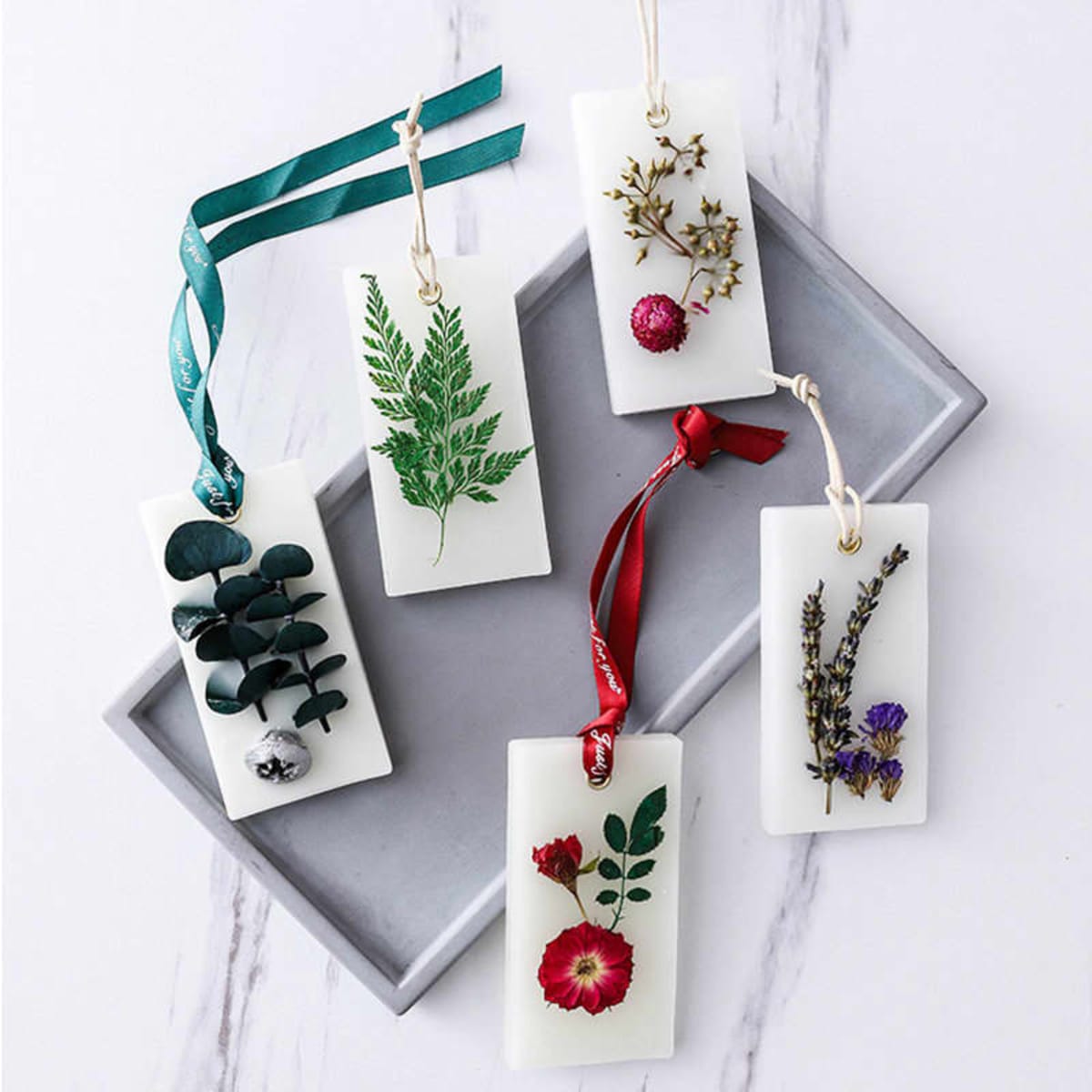 A set of five dried flower scented wax tablets in various designs, displayed on a grey tray with colourful ribbons.