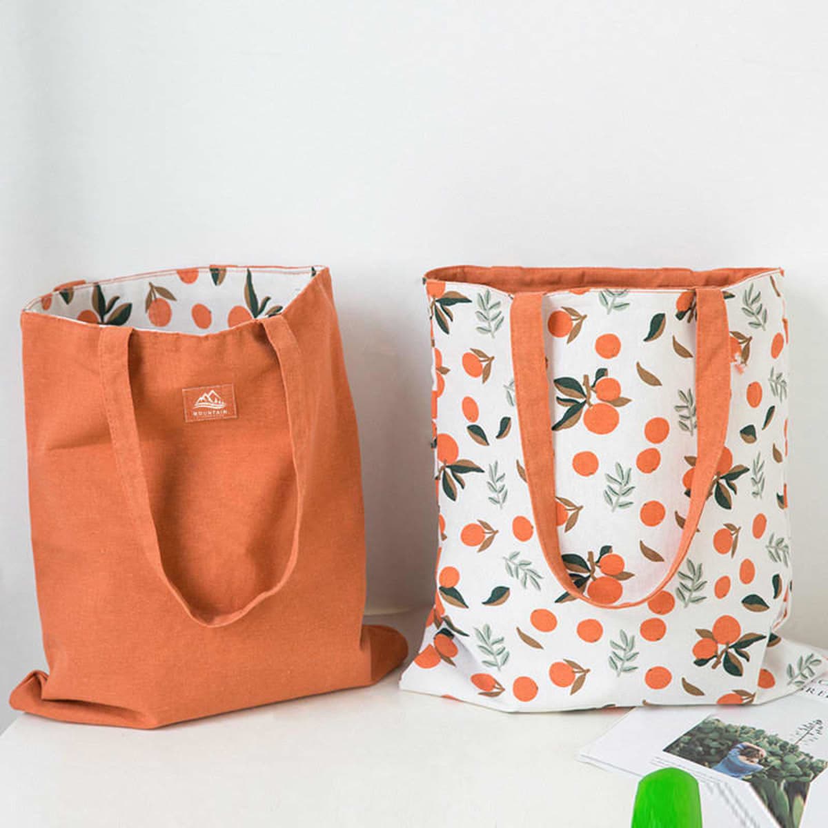 Two reusable canvas grocery bags in orange and a white design featuring orange fruit patterns, each with sturdy handles.