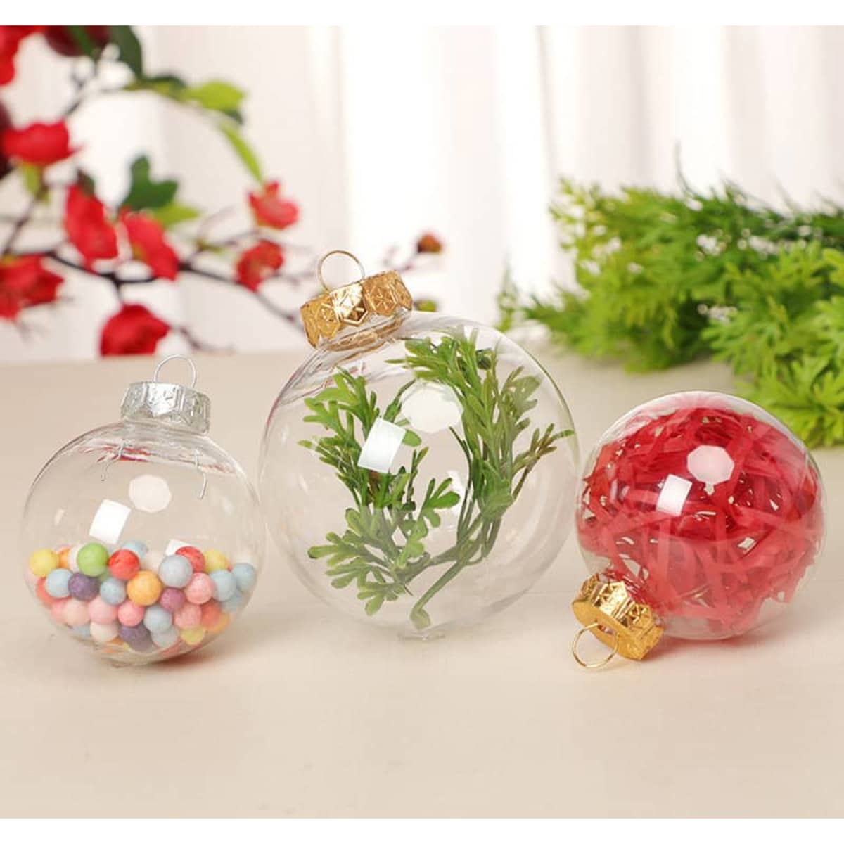 Three clear plastic ball ornaments in varying sizes filled with colourful beads, green foliage, and red filler, featuring gold tops.