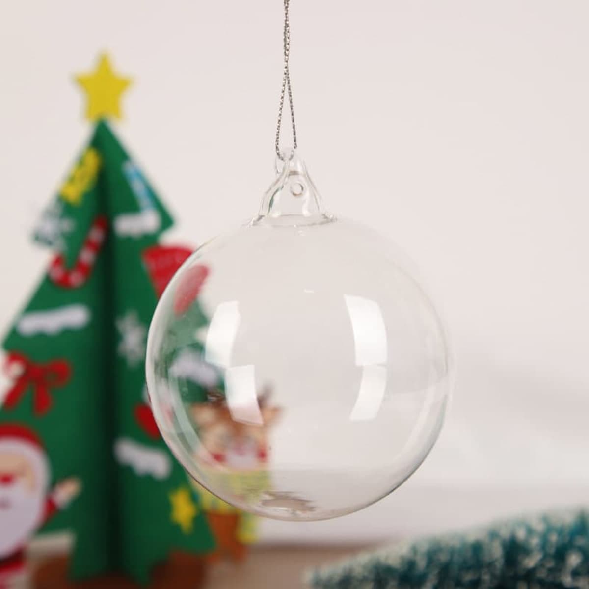 A clear 6cm transparent glass pendant ball, featuring a hanging loop, against a festive background of a decorated Christmas tree.