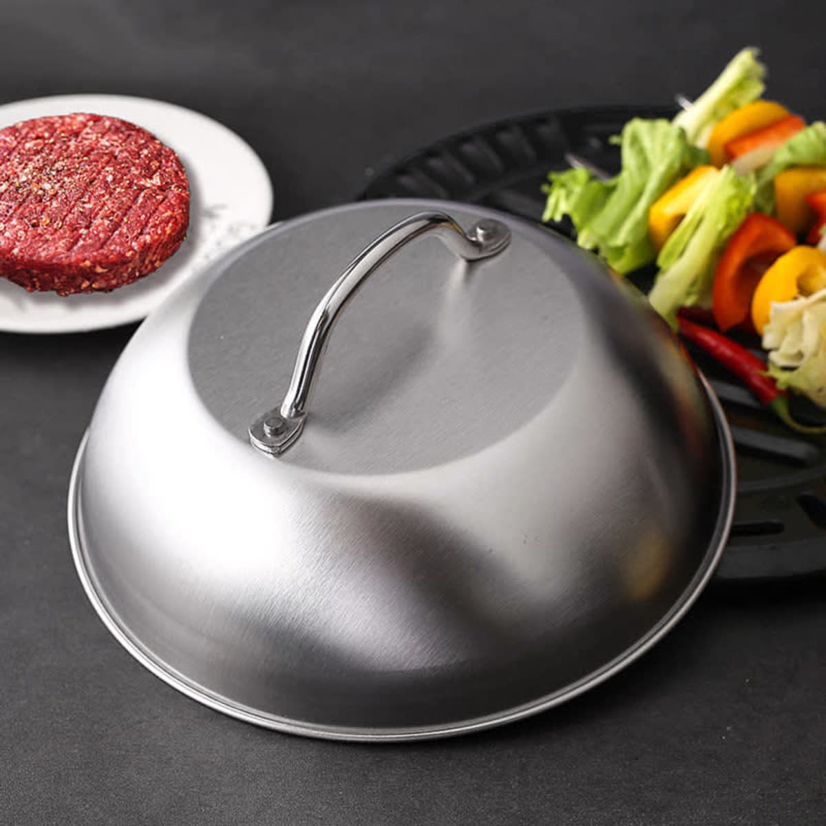 A 9-inch round stainless steel basting cover with a handle, placed next to a plate of vegetables and a burger patty.