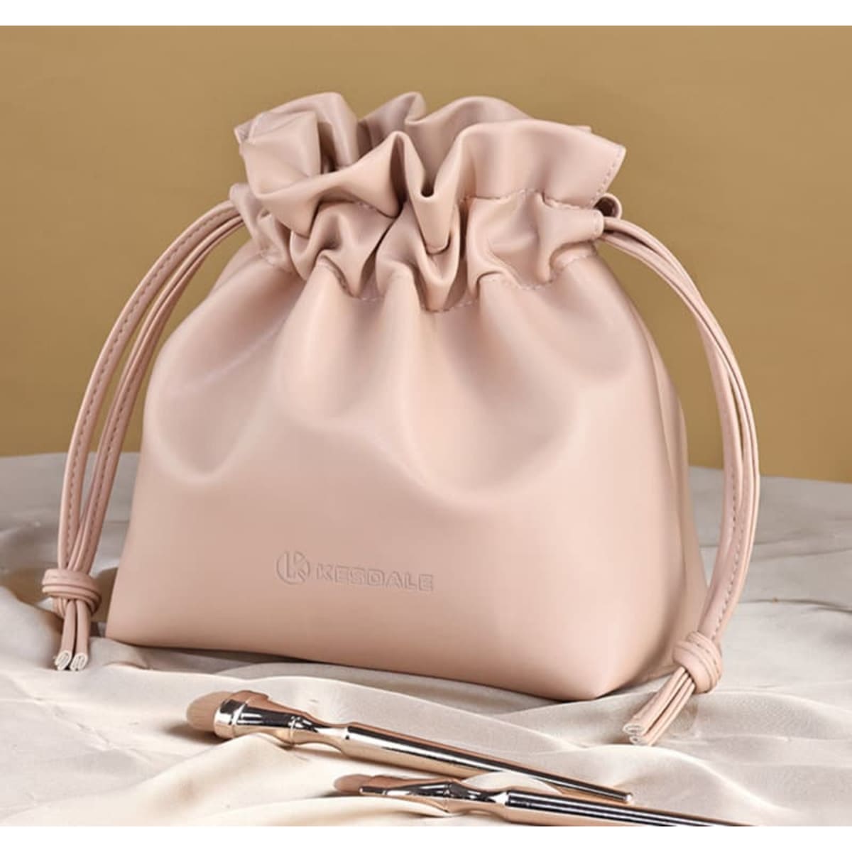 A pink drawstring make-up bag with a ruffled top, accompanied by two make-up brushes. It features a logo design.