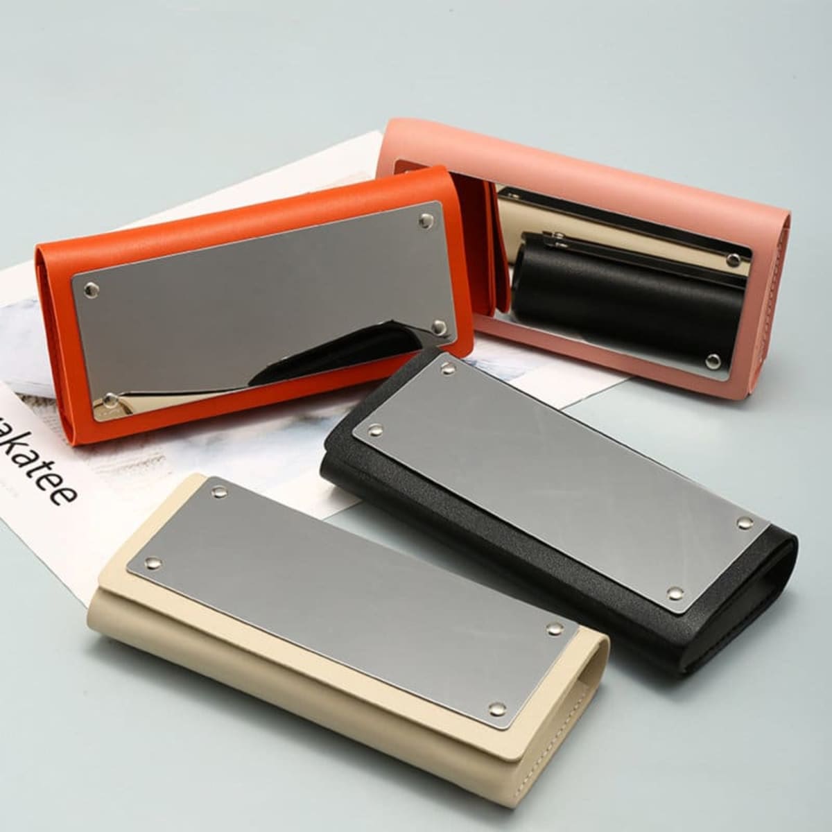 A selection of spectacle cases in orange, pink, black, and beige, each featuring a mirror on the front.