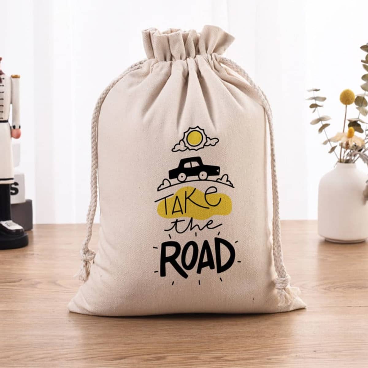 A cream cotton drawstring storage bag featuring a graphic with a car, sun, and clouds. It has a branding logo.