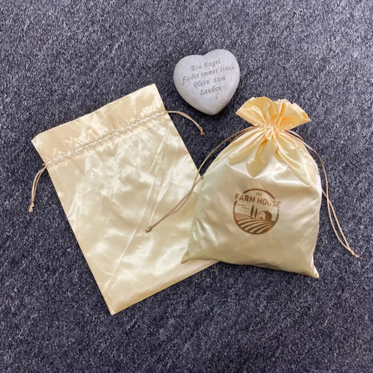 A gold satin drawstring bag and a plain satin bag, both featuring a logo, alongside a decorative stone.