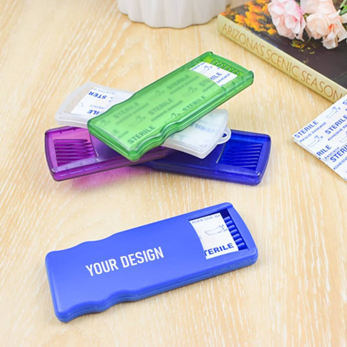 Five adhesive bandage containers in purple, green, and blue, stacked on a wooden surface with floral decor.