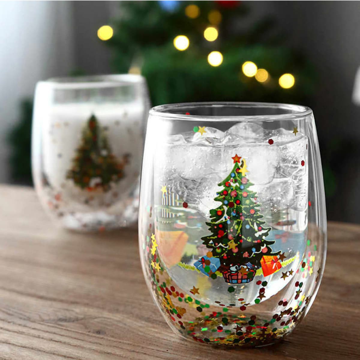 A decorative double wall glass mug featuring a Christmas tree design with colourful accents, set against a blurred festive background.