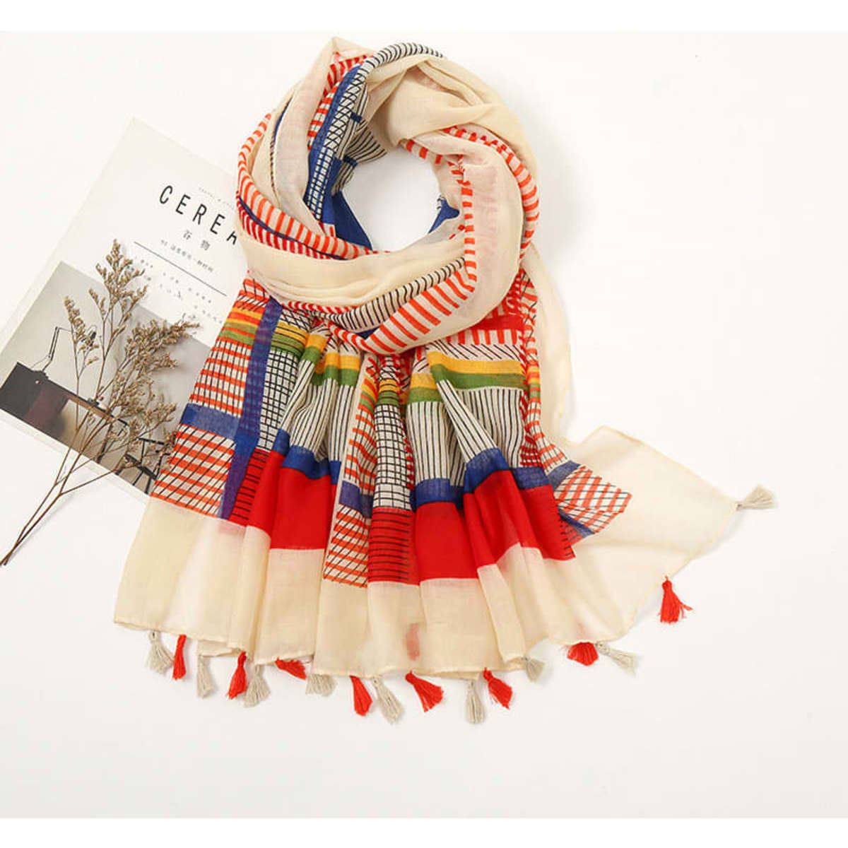A multi-coloured cotton scarf featuring red, blue, and cream patterns with tassels at the ends and a floral backdrop.