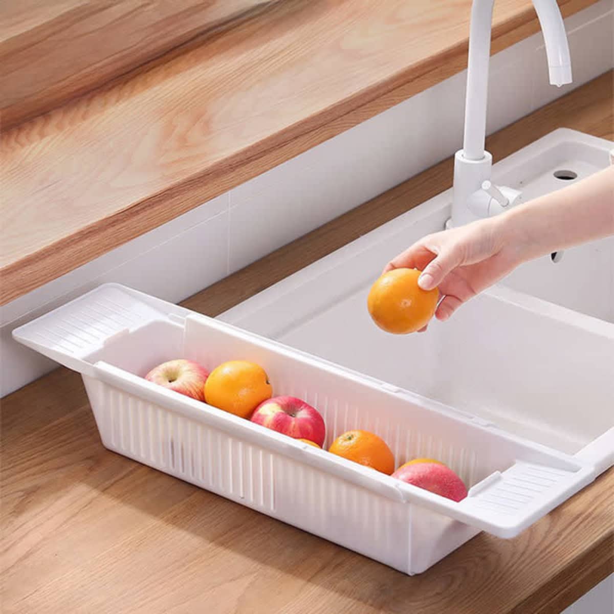 A white rectangular sink basket holding various fruits including apples and oranges, positioned next to a sink.