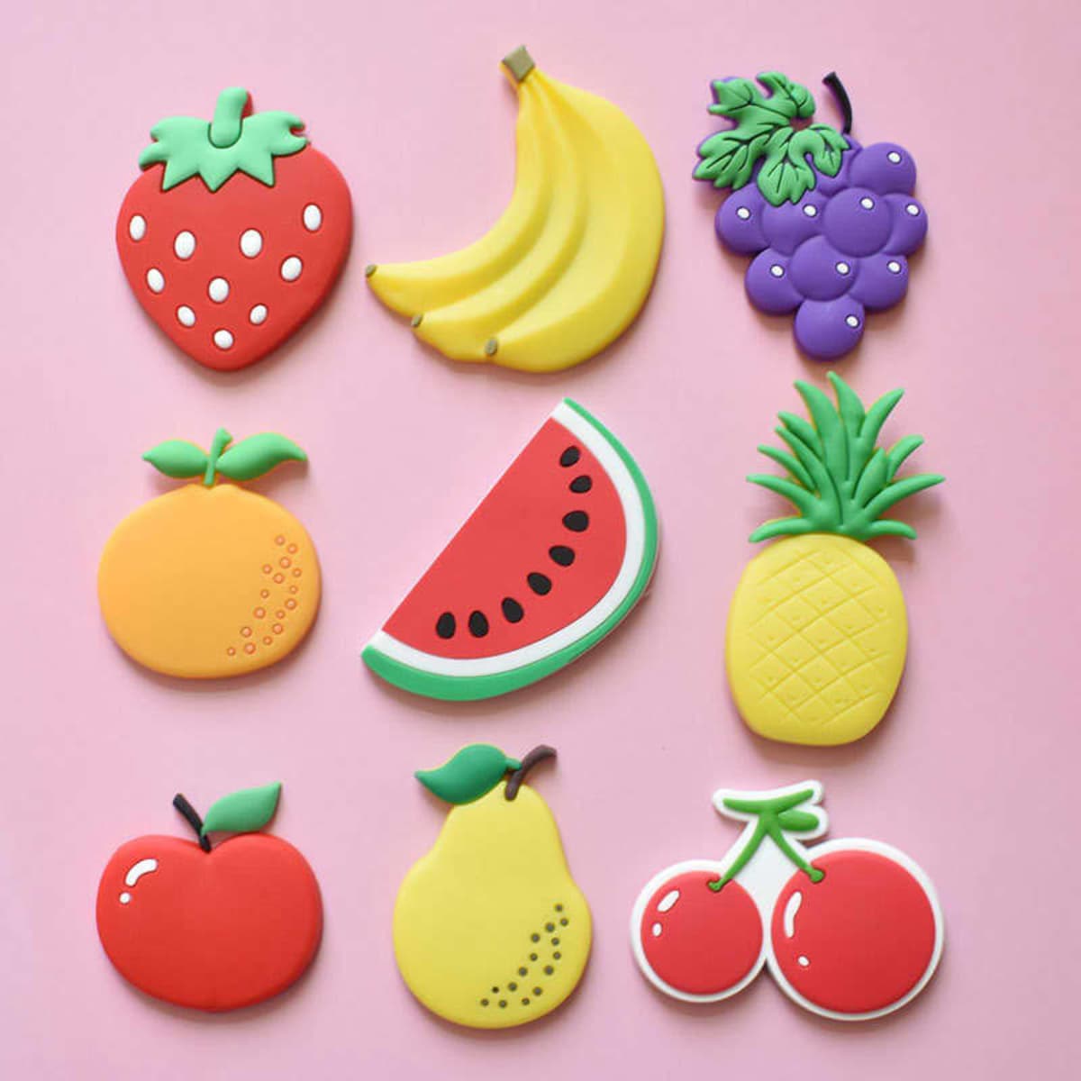 A collection of colourful fruit-shaped fridge magnets, including strawberries, bananas, grapes, watermelon, and others on a pink background.