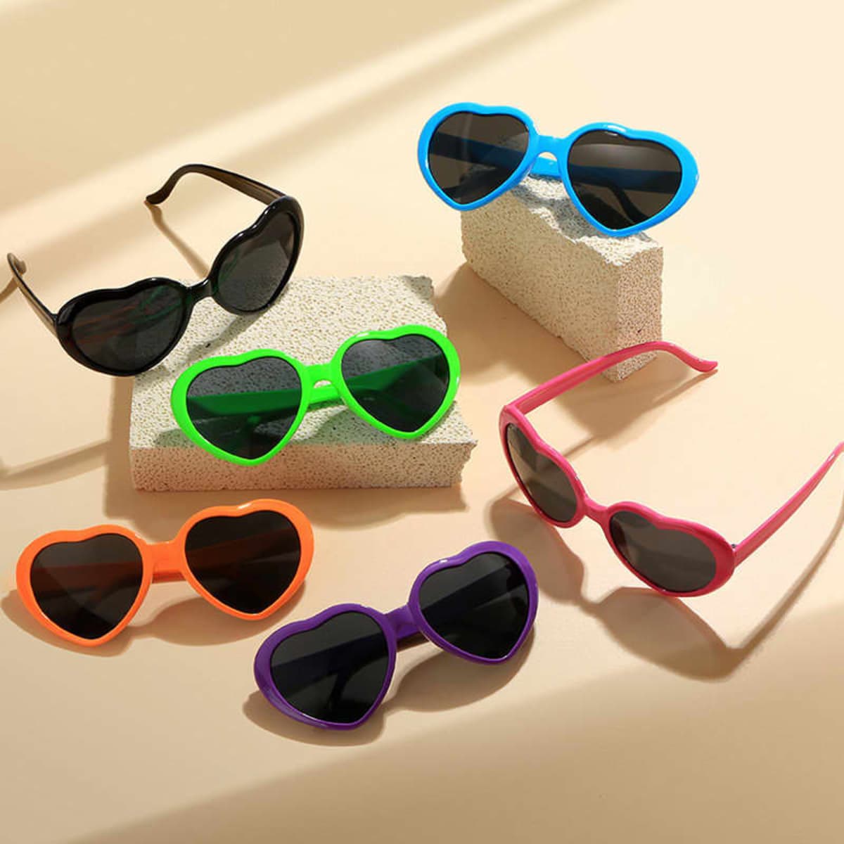 A variety of heart-shaped sunglasses in black, blue, green, orange, pink, and purple displayed on light-coloured surfaces.