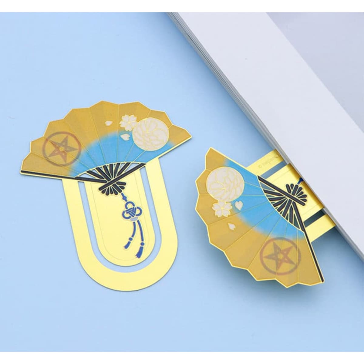 A golden fan-shaped metal bookmark with intricate blue and white designs, placed beside a book.