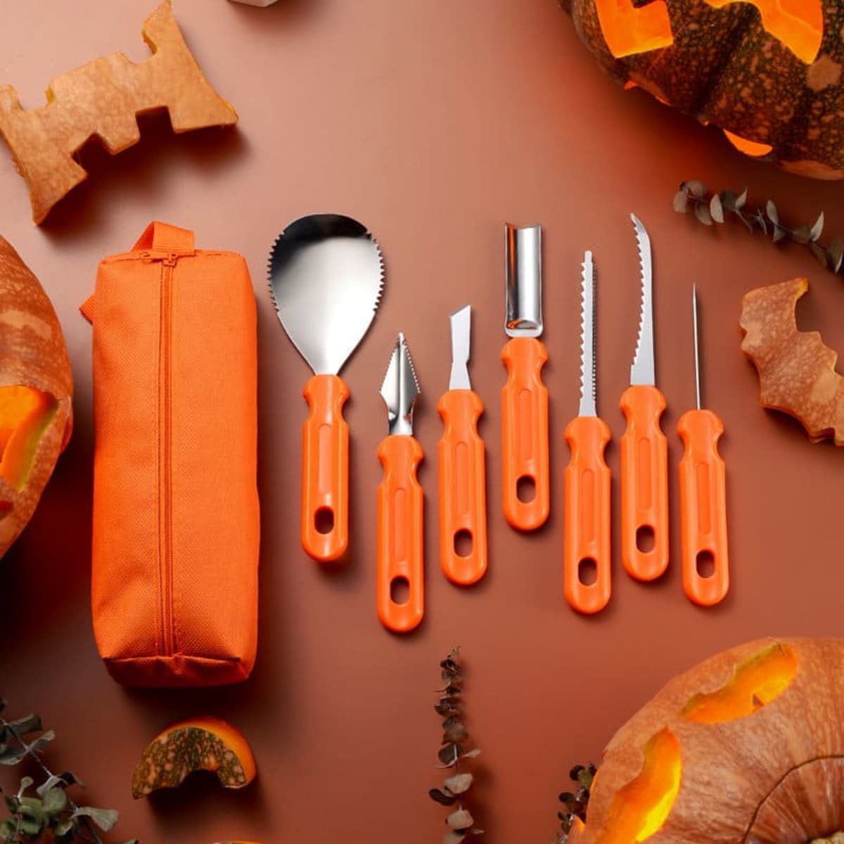 A set of seven pumpkin carving tools with orange handles, displayed with carved pumpkins and a carrying pouch.