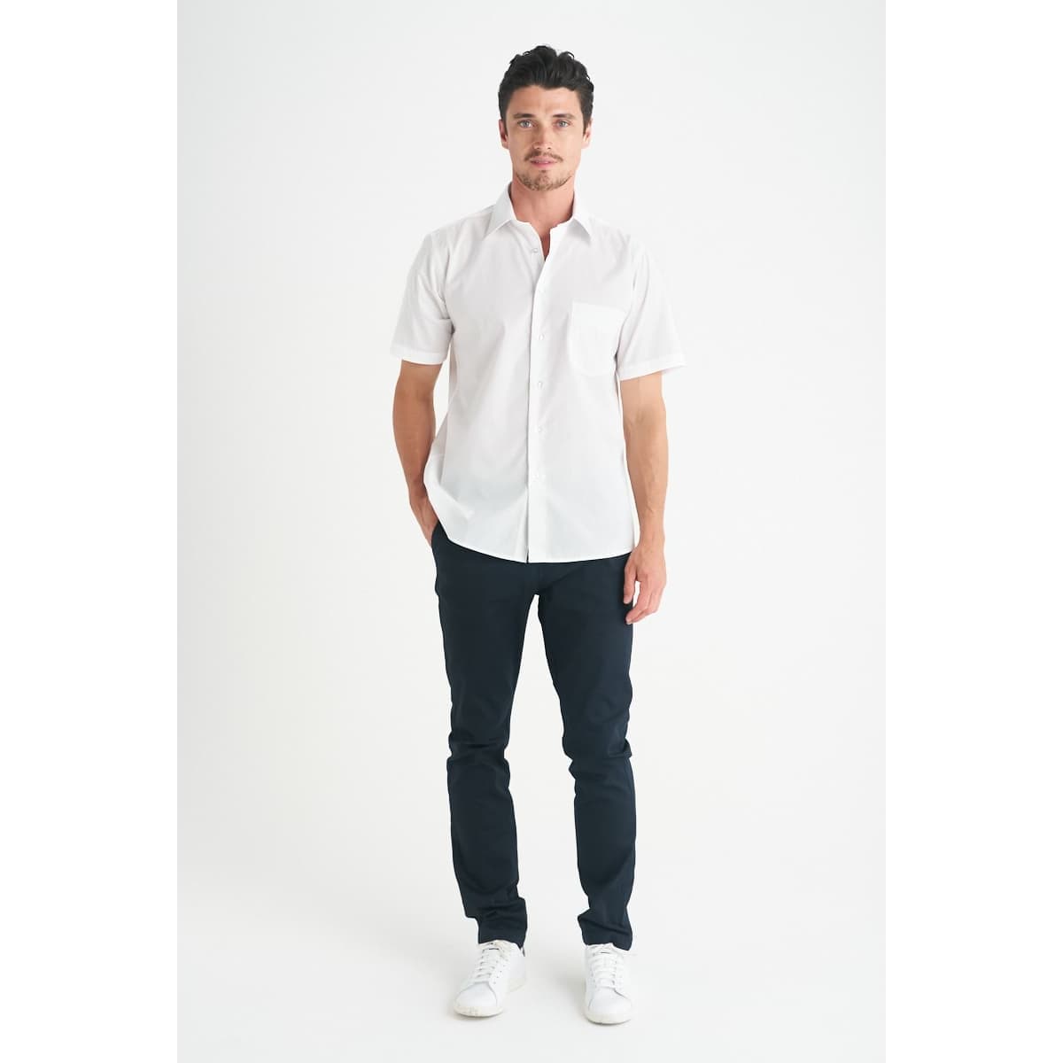 A man wearing a white short sleeve button-up shirt with a chest pocket, paired with dark trousers and white shoes.