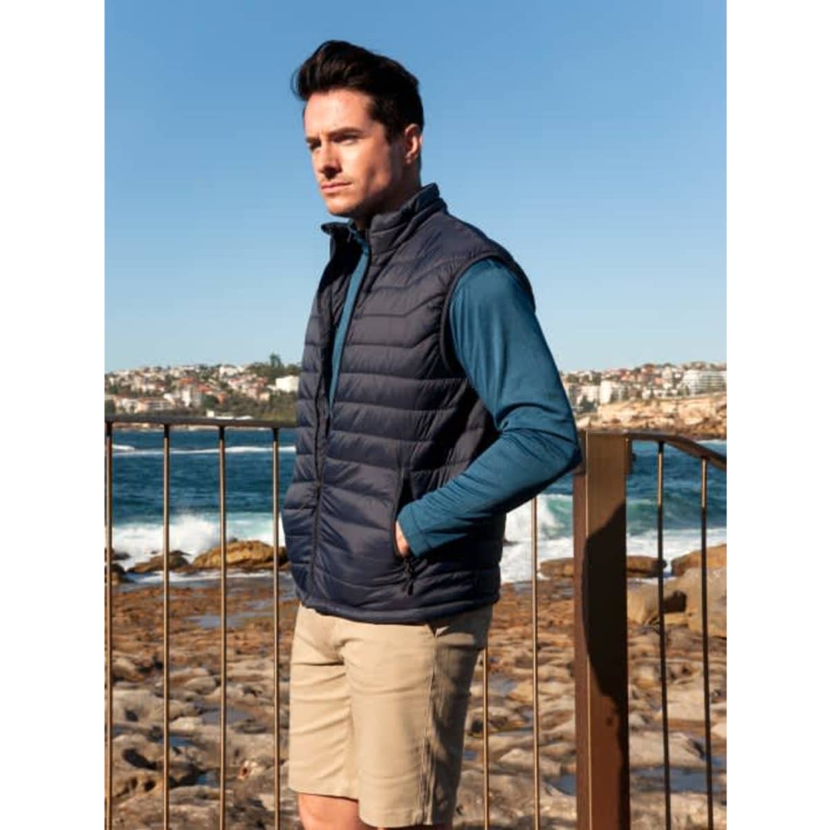 A navy puffer vest worn over a long-sleeve blue top, with a scenic coastal background. The model stands by a railing.