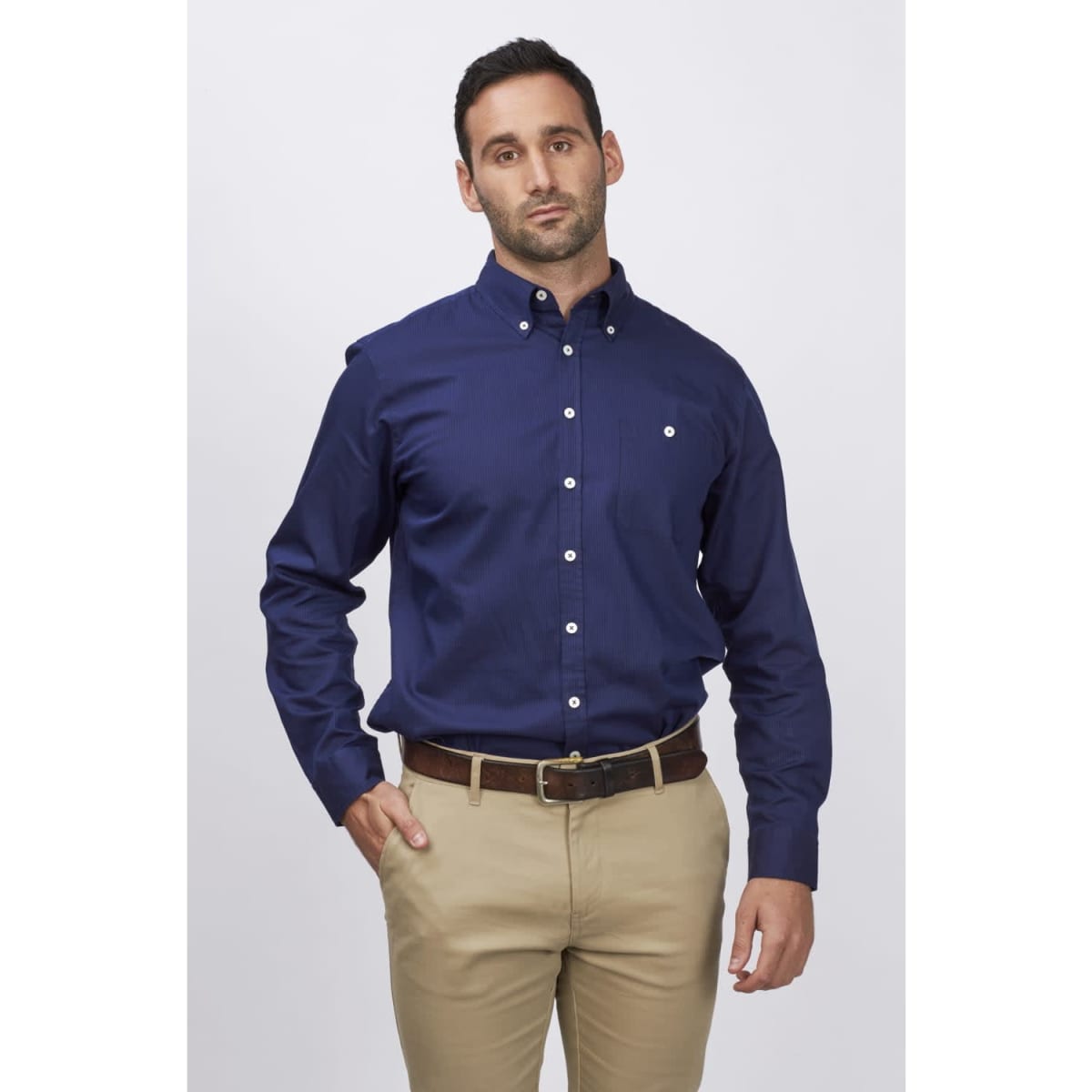 A men's long sleeve button-up shirt in navy blue, featuring a chest pocket and a logo. Paired with light-coloured trousers.