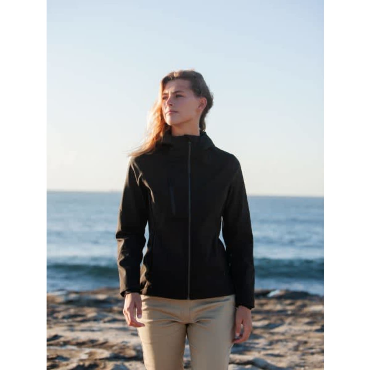 A woman stands by the beach in a black soft shell jacket, featuring a zip front and a sleek design.
