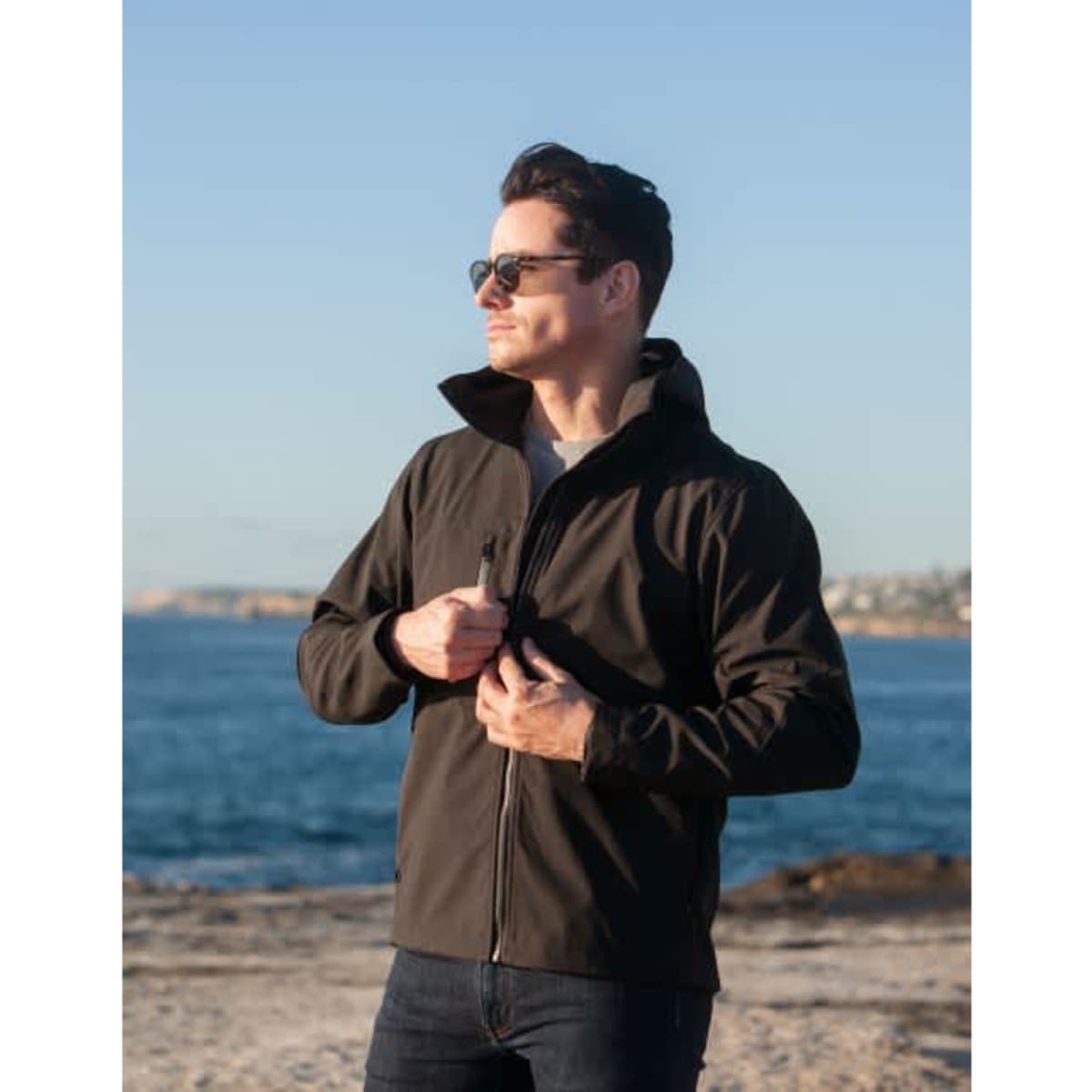 A man wearing a black soft shell jacket stands by the water, adjusting his jacket with sunglasses on.