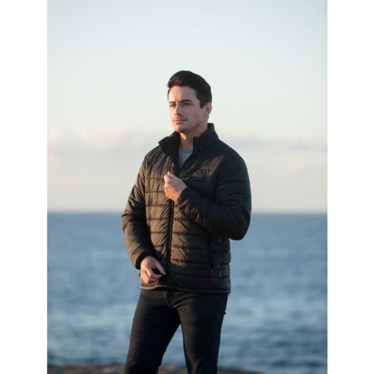 A black puffer jacket for men, featuring a zip-up front and a logo, worn by a model against a coastal backdrop.
