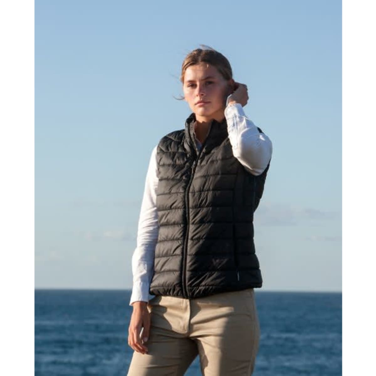 A woman stands outdoors wearing a black puffer vest over a white shirt, with the ocean in the background.