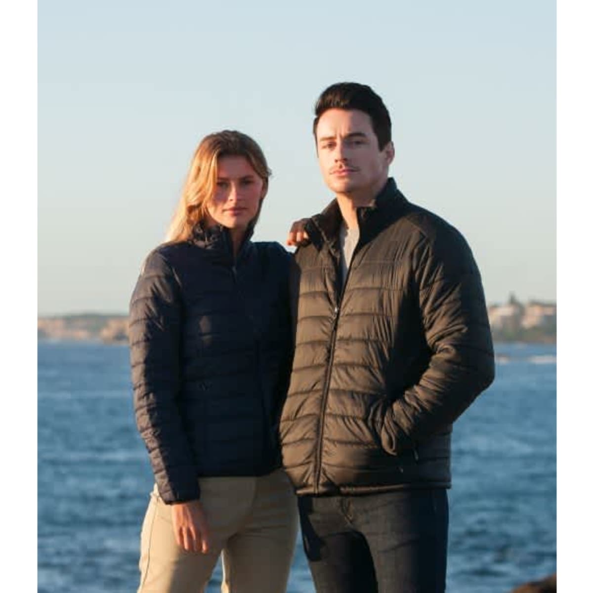 A couple stands by the water wearing navy puffer jackets. The scene is coastal with a clear sky.