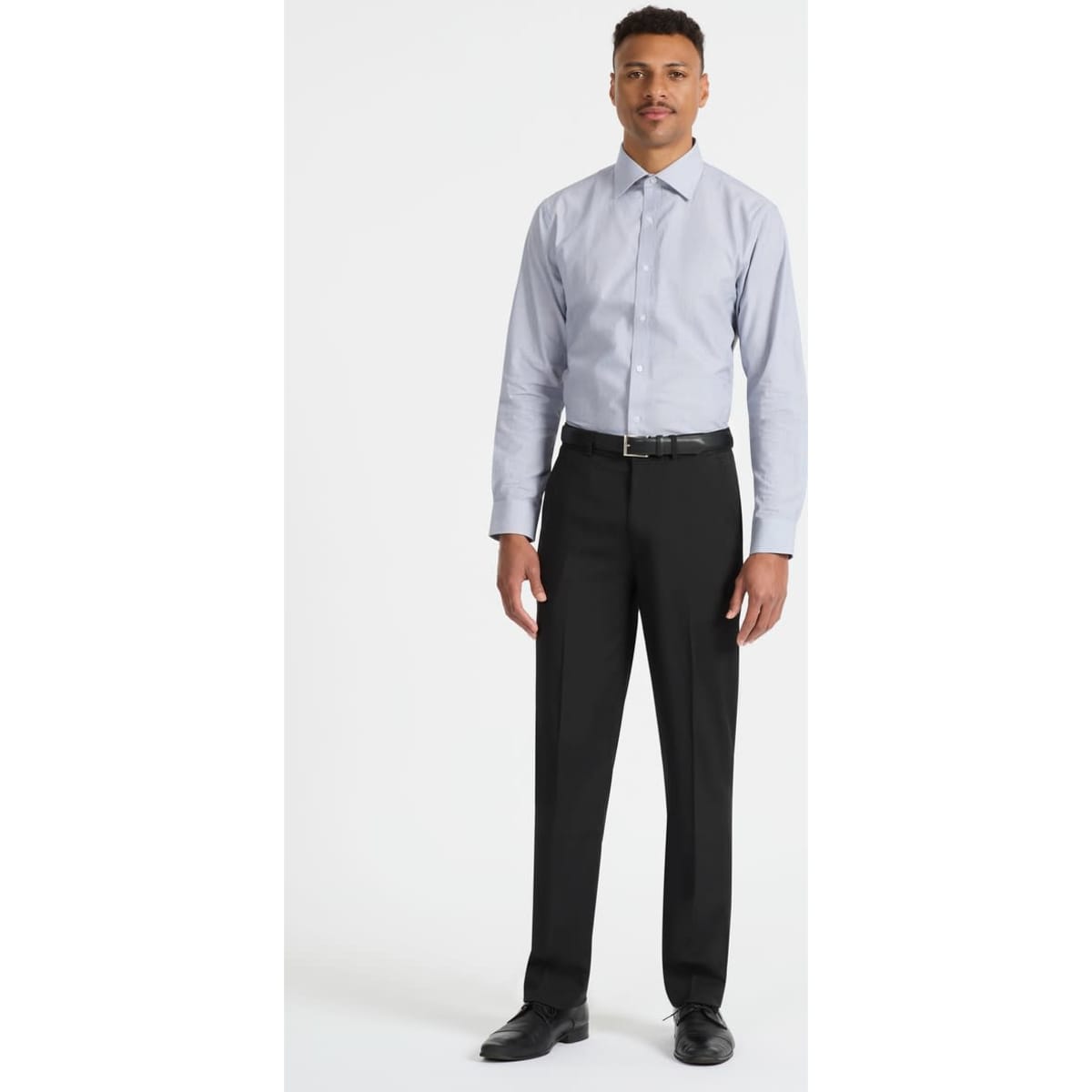 A man wears a light blue long sleeve button-up shirt paired with black trousers and shoes. The shirt features a logo.