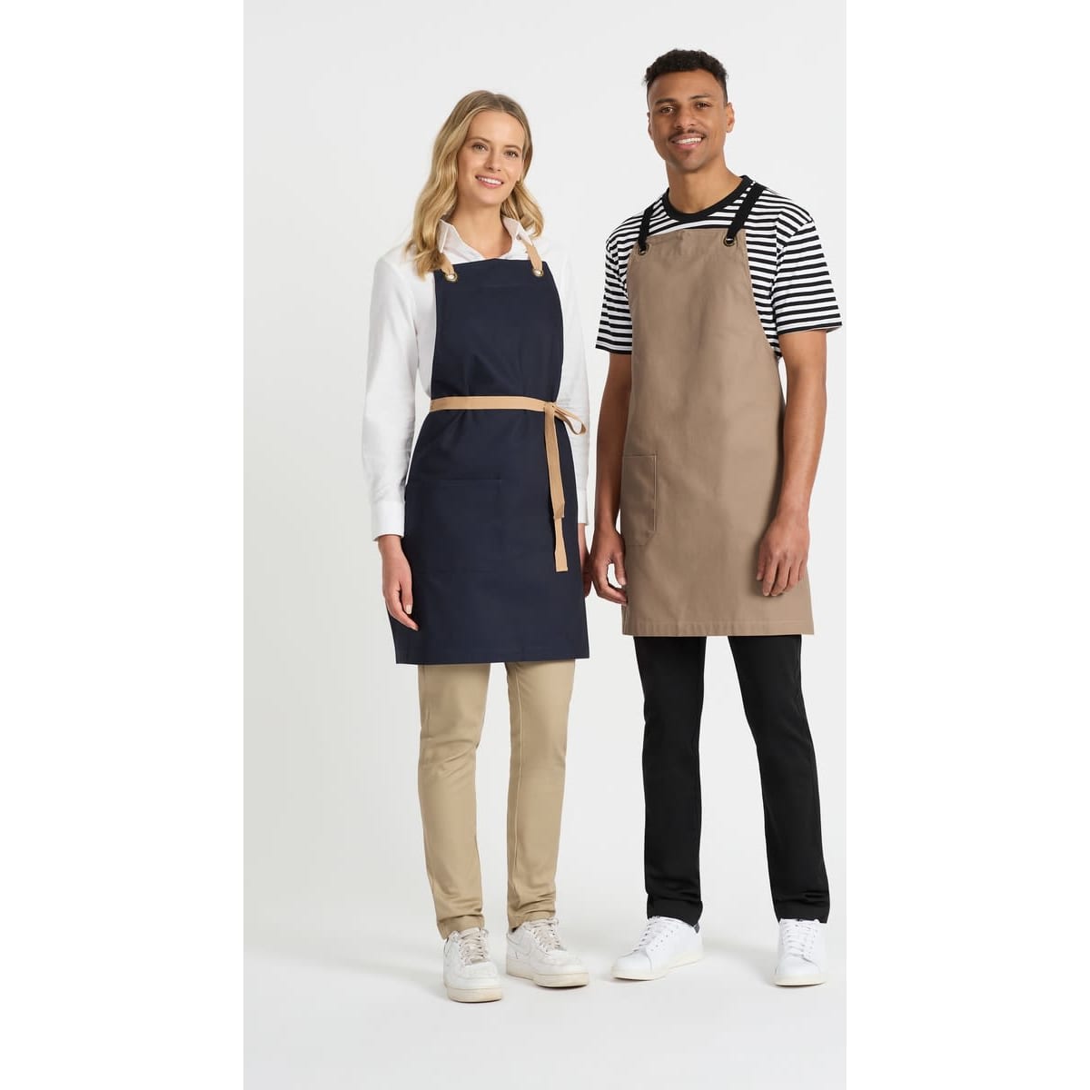 A navy bib apron on a woman and a beige bib apron on a man, both with shoulder straps, standing together.