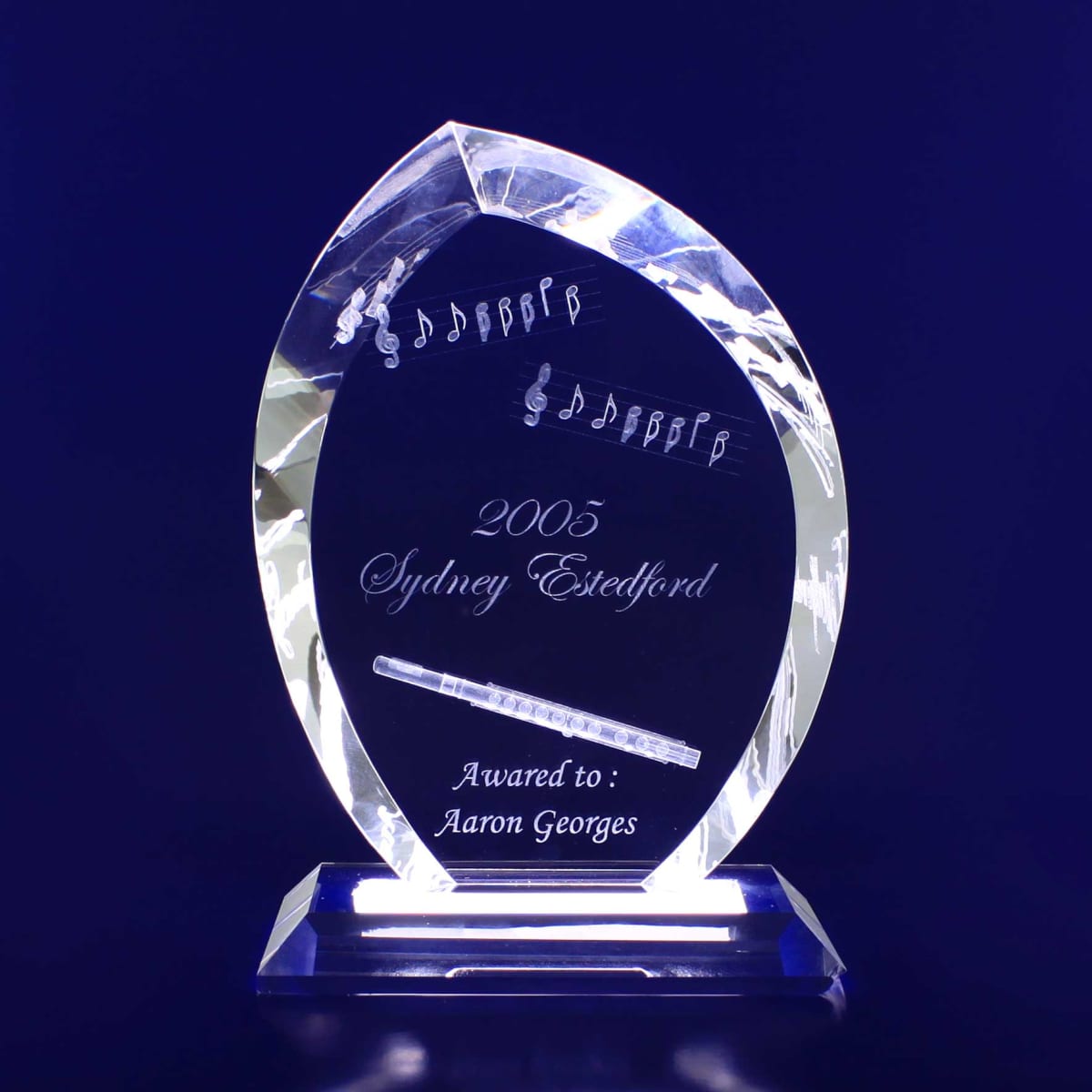 A crystal trophy shaped like a flame, engraved with musical notes and a pen design, on a clear base.