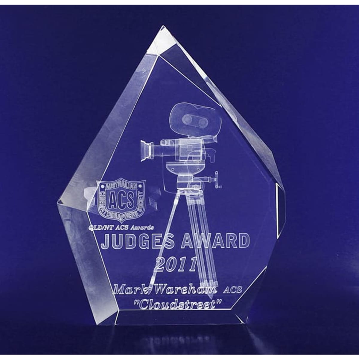 A clear, crystal trophy with a geometric design, featuring an engraved film camera and text, on a dark background.