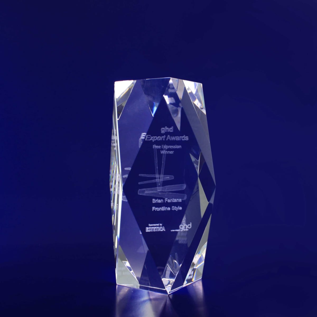 A clear crystal award with a bevelled edge, featuring an engraved design and logo, set against a dark blue background.
