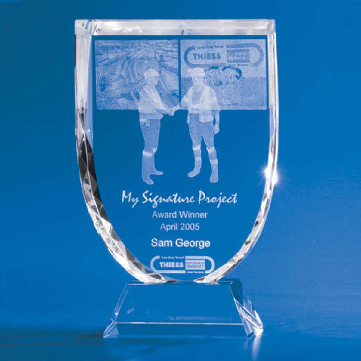 A crystal trophy with a shield shape, featuring two figures shaking hands, set against a blue background.