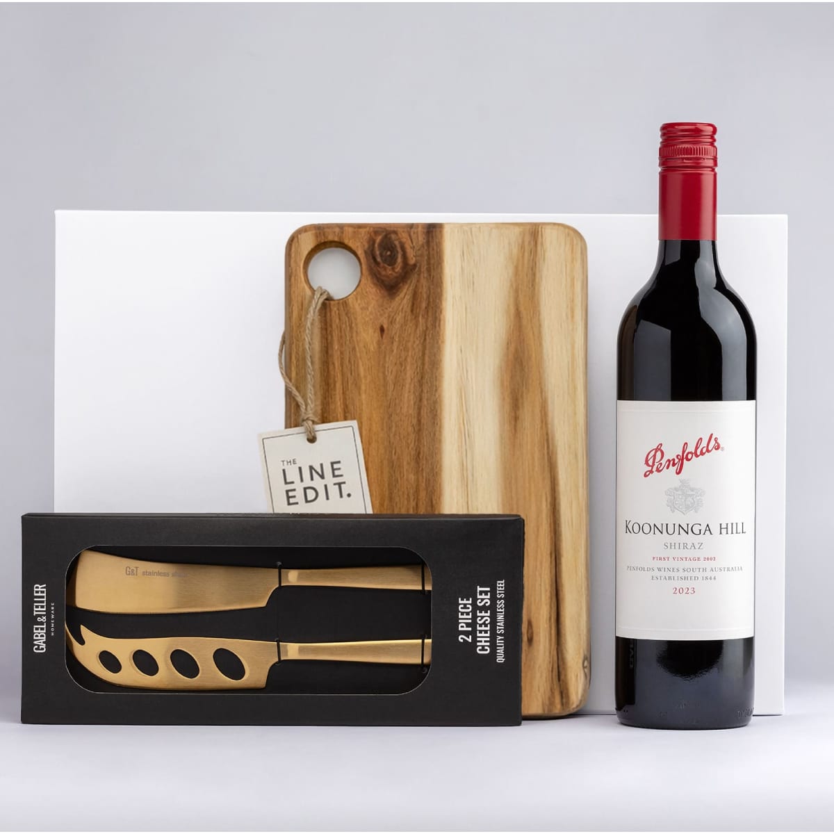 A wooden cheese board with a knife set in a box and a bottle of red wine. The board has a rounded cutout.