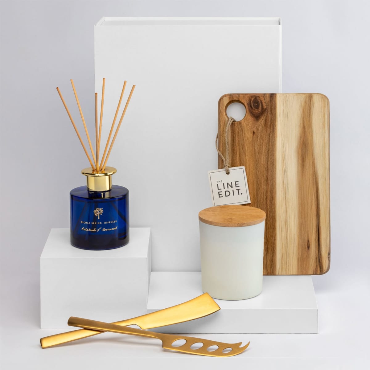A blue diffuser with wooden sticks, a white candle in a ceramic container, and a wooden cheese board with gold knives.