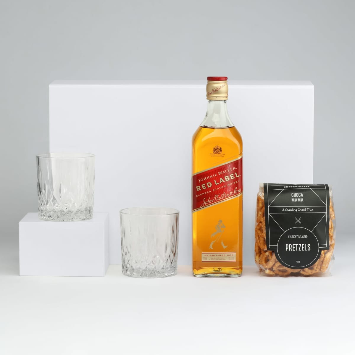 A drink bottle of Johnnie Walker Red Label whisky, two crystal glasses, and a pack of pretzels on a neutral background.